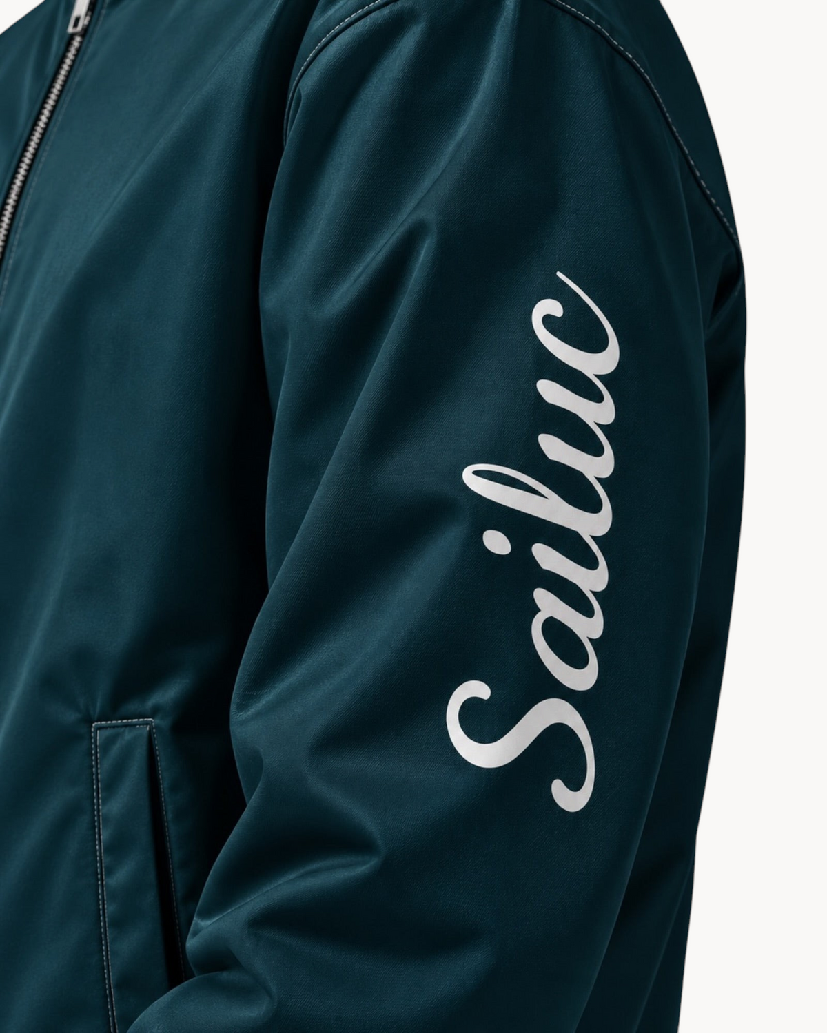 Close-up of a dark teal Sailuc Vesper bomber jacket featuring a white script logo on the sleeve and silver zipper.