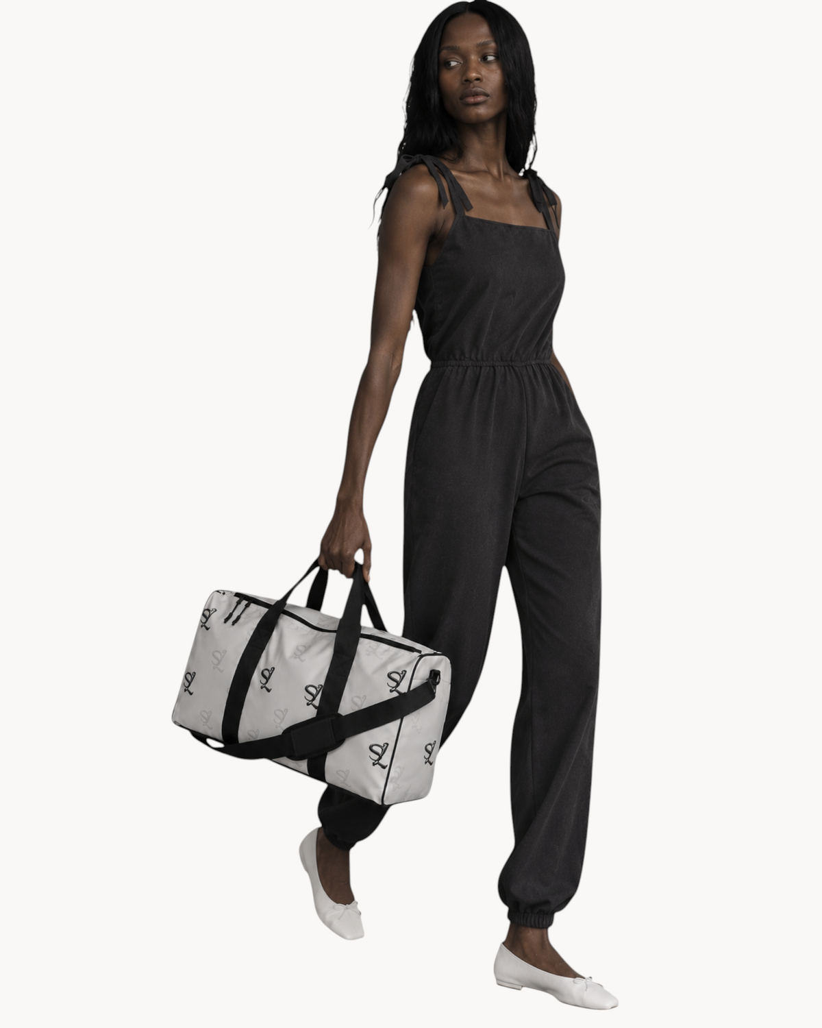 A model carrying a white Sailuc Essential Voyage Bag with a black monogram print and black handles in a studio.