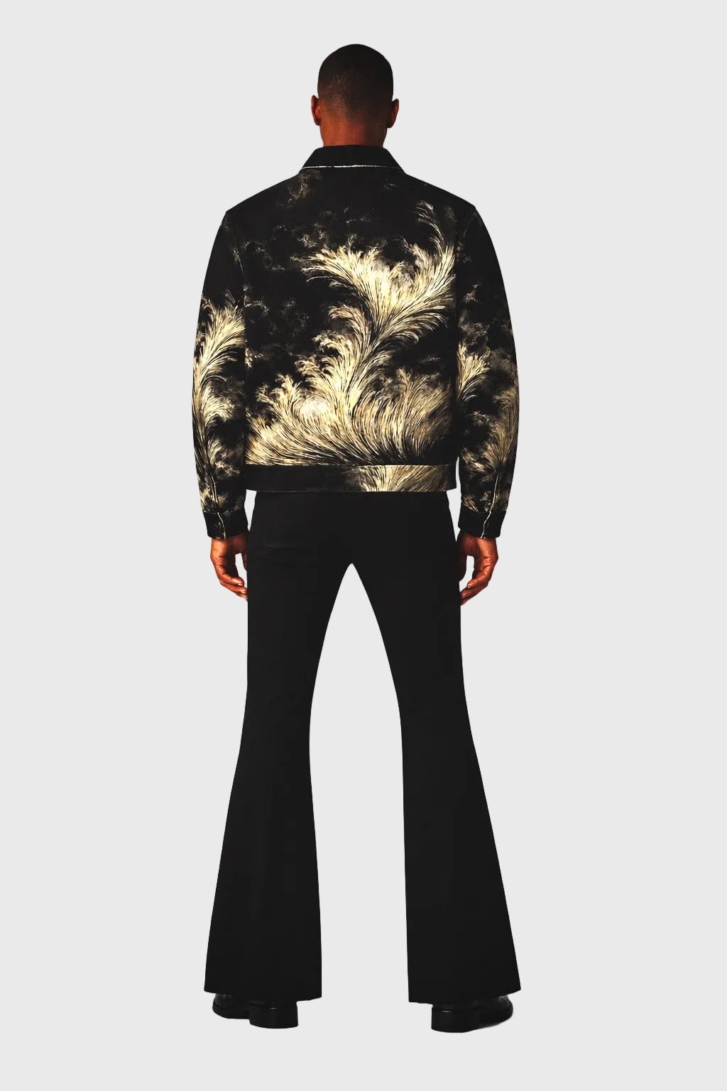 Person wearing a black feather-patterned jacket with black bell-bottom pants on a gray background