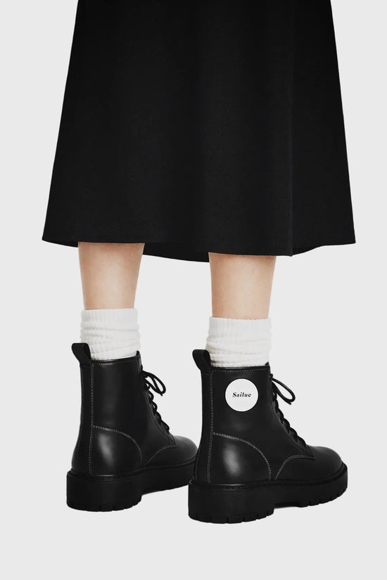 Person wearing black boots with a visible brand logo, white socks, and a black skirt on a gray background.