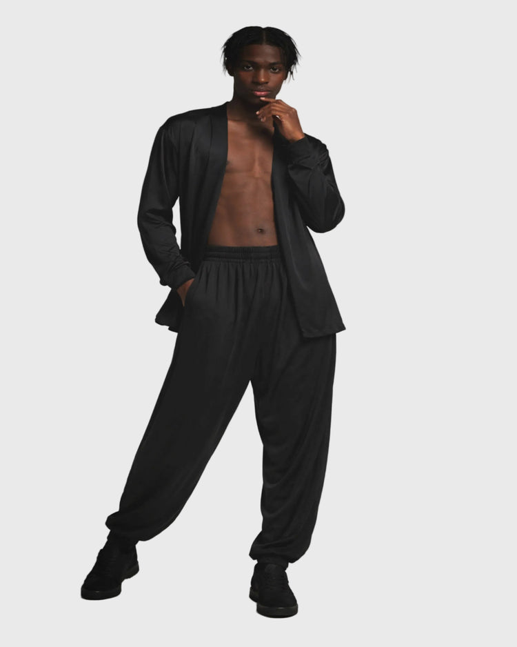 Man wearing a black cardigan over loose fitted trousers against a gray background